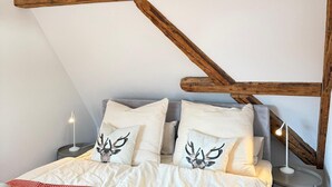 Un dormitorio con techo inclinado, vigas de madera a la vista, cama con ropa blanca y almohadas estampadas, manta de diseño rojo y blanco, y lámparas de noche.