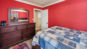 A bedroom with a red wall, a wooden dresser, a bed with a quilt, a mirror, and a door leading to another room. - Chipmunk House (Big Bear Lake)