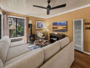 A living room with a sofa, a fireplace, a ceiling fan, a painting, and a window with curtains. - Chipmunk House (Big Bear Lake)