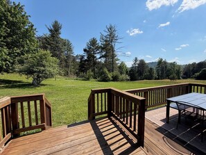 Una terraza de madera con mesa y sillas, rodeada por un campo cubierto de hierba y árboles.