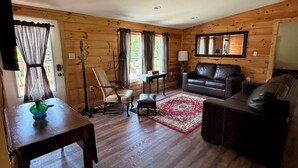 A cozy living room with a wooden floor, a black leather sofa, a dining table with chairs, and a red rug. - Cabin Fever II is a cozy 3-bedroom cabin in the foothills of the Blue Ridge.  (Luray)