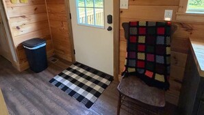 A wooden interior with a door, a window, a trash bin, a checkered mat, and a chair with a colorful blanket. - Cabin Fever II is a cozy 3-bedroom cabin in the foothills of the Blue Ridge.  (Luray)