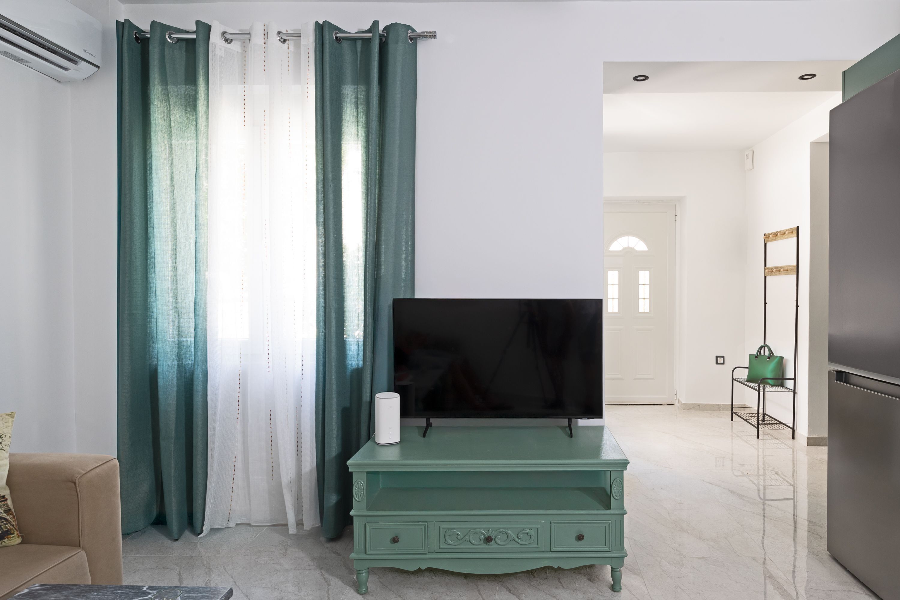 A modern living room with a flat-screen TV on a teal console, a beige sofa, and a green handbag on a shelf.