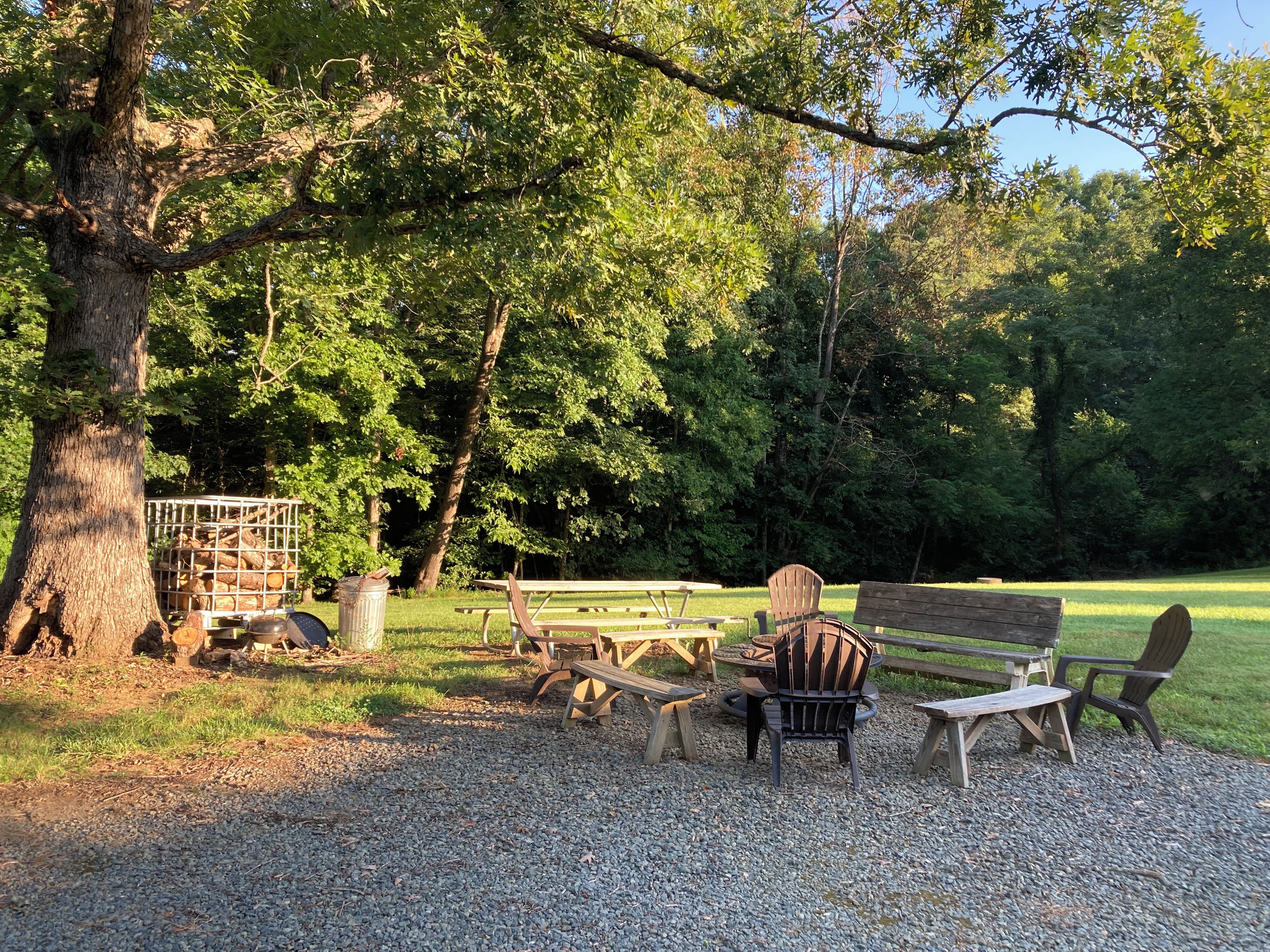 A picnic area with wooden benches and chairs, surrounded by trees and a gravel ground.