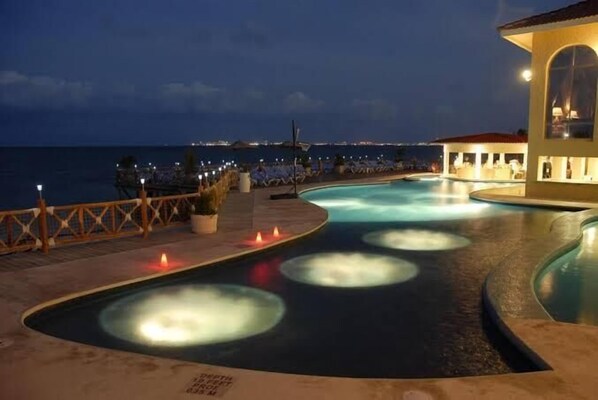 A pool area with a view of the ocean at night, a lounge area with chairs, and a building with a red roof. - tropical apartment (Cancún)