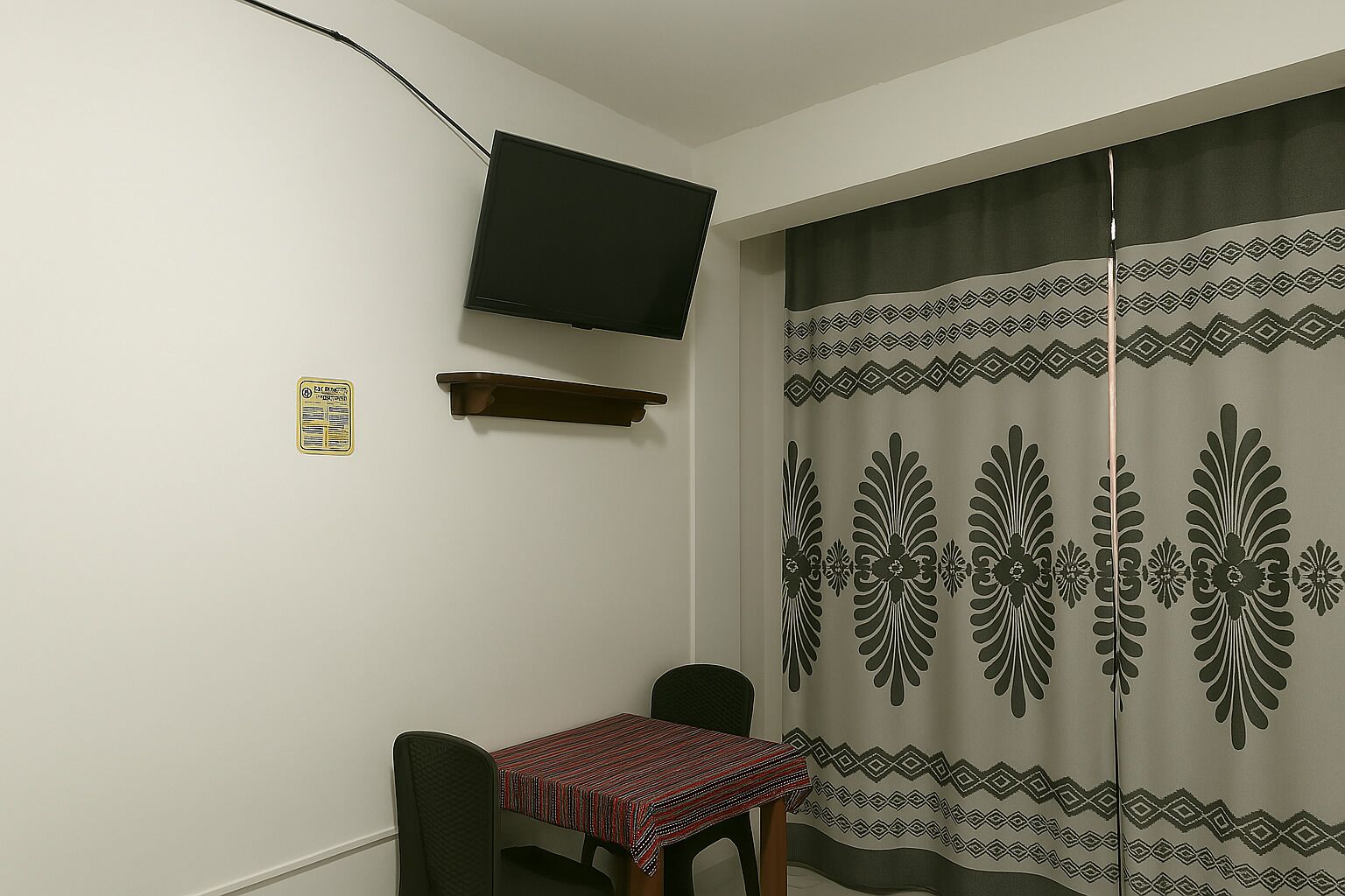 A small room with a TV mounted on the wall, a table with a patterned tablecloth, and a curtain with a geometric design.