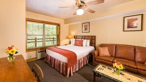A hotel room with a bed, a brown leather sofa, a wooden coffee table, a dining table with chairs, and a ceiling fan.