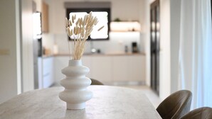 Un jarrón blanco con plantas secas sobre una mesa de mármol en una cocina moderna con armarios blancos y un ventanal.
