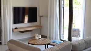 Una sala de estar moderna con una televisión de pantalla plana montada en la pared, una mesa de centro de madera, un sofá color beige y vistas al exterior a través de amplios ventanales.
