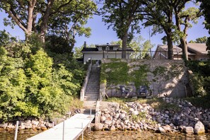 Une maison avec un escalier en pierre menant à une terrasse, entourée d’arbres et bordée d’une côte rocheuse.