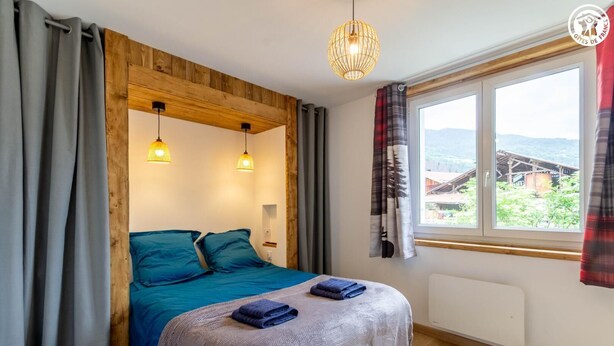 A bedroom with a wooden ceiling, two hanging light fixtures, a bed with blue bedding, a window with a view of a building, and a radiator. - MUTCHOS - LA PLAGNE TARENTAISE (LA PLAGNE TARENTAISE)