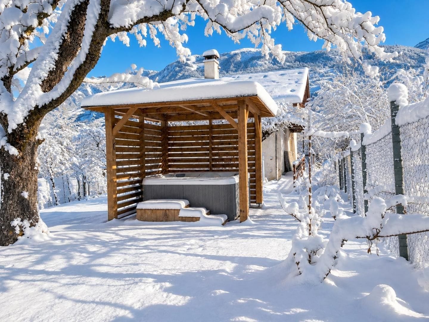 A wooden outdoor hot tub area with a wooden pergola structure, surrounded by snow-covered trees and a mountain backdrop.