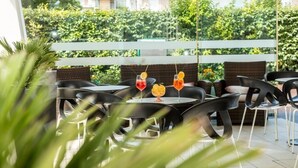 An outdoor seating area with round tables and black chairs, surrounded by greenery and a white fence. - Carinthia (Lido di Jesolo)