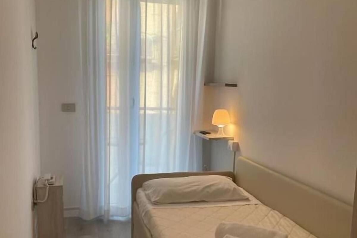 Un dormitorio pequeño y ordenado con una cama, una lámpara de noche y una ventana con cortinas traslúcidas.