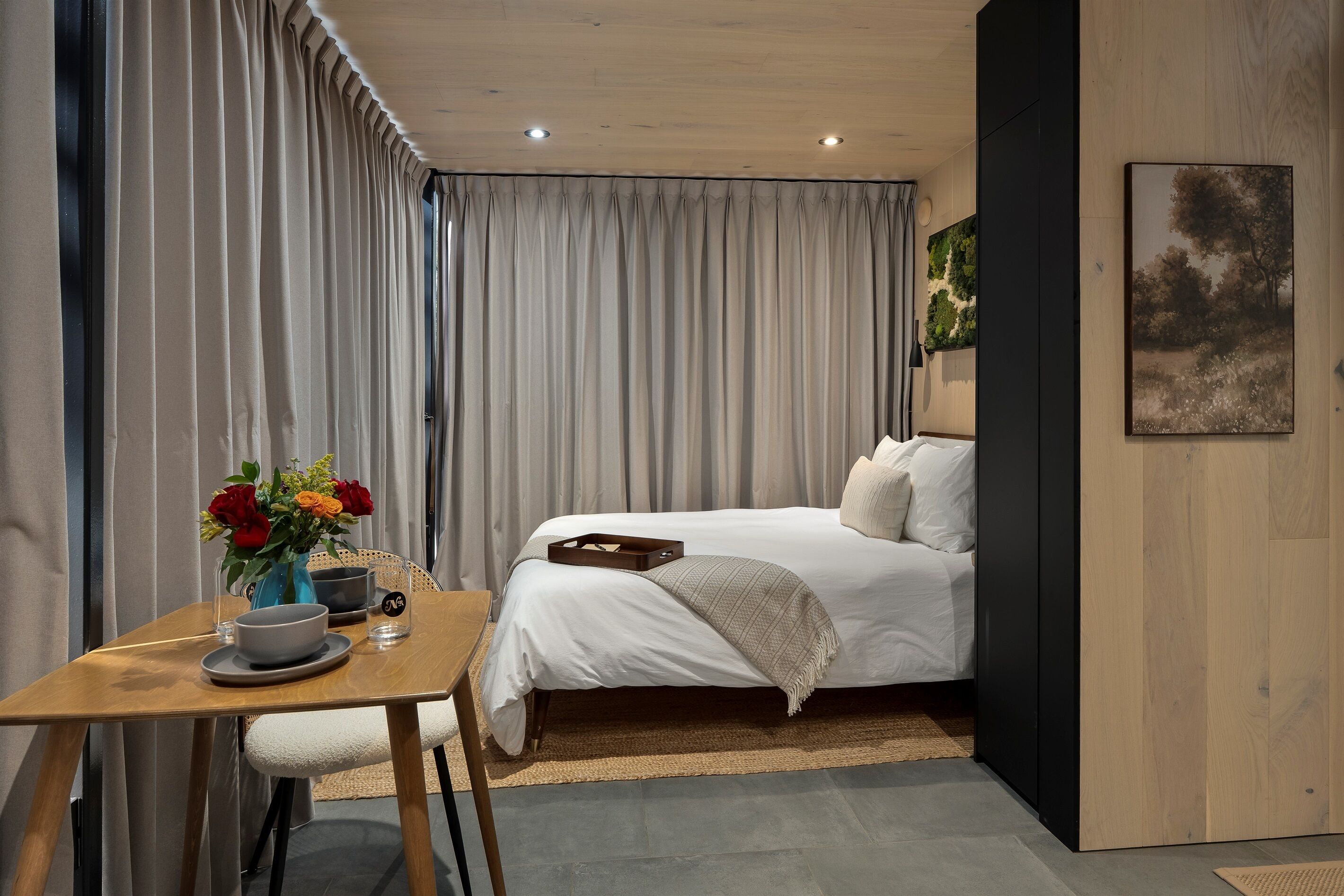 Ein modernes Hotelzimmer mit einem Bett, einem Holztisch mit Blumenvase und einem Bild an der Wand.