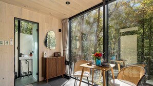 A modern room with a large window, a wooden door, a round mirror, a wooden cabinet, a sink, and a table with chairs. - Mirrored Glass Studio · Hot Tub · Nature Views (Laurelville)