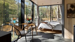 A modern bedroom with a large window, a bed, a table with chairs, and a view of a forest. - Mirrored Glass Studio · Hot Tub · Nature Views (Laurelville)