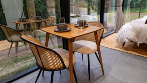 A dining area with a table set for two, surrounded by wicker chairs, and a bed with a white duvet in the background. - Mirrored Glass Studio · Hot Tub · Nature Views (Laurelville)