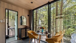 Een modern interieur met een glazen tafel, twee gele stoelen, een ronde spiegel en uitzicht op een bos door grote ramen.