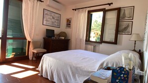 Ein Schlafzimmer mit einem Bett, einem Schreibtisch mit Computer, einem Stuhl, einem Fenster mit Vorhängen und einer Heizung.
