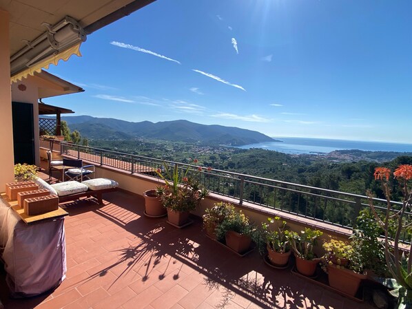 A balcony with outdoor furniture, potted plants, and a scenic view of mountains and a body of water.