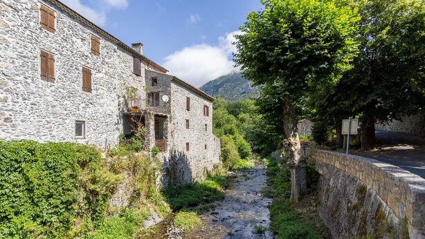 A stone building with multiple windows and a balcony, a narrow stream, and a stone wall.