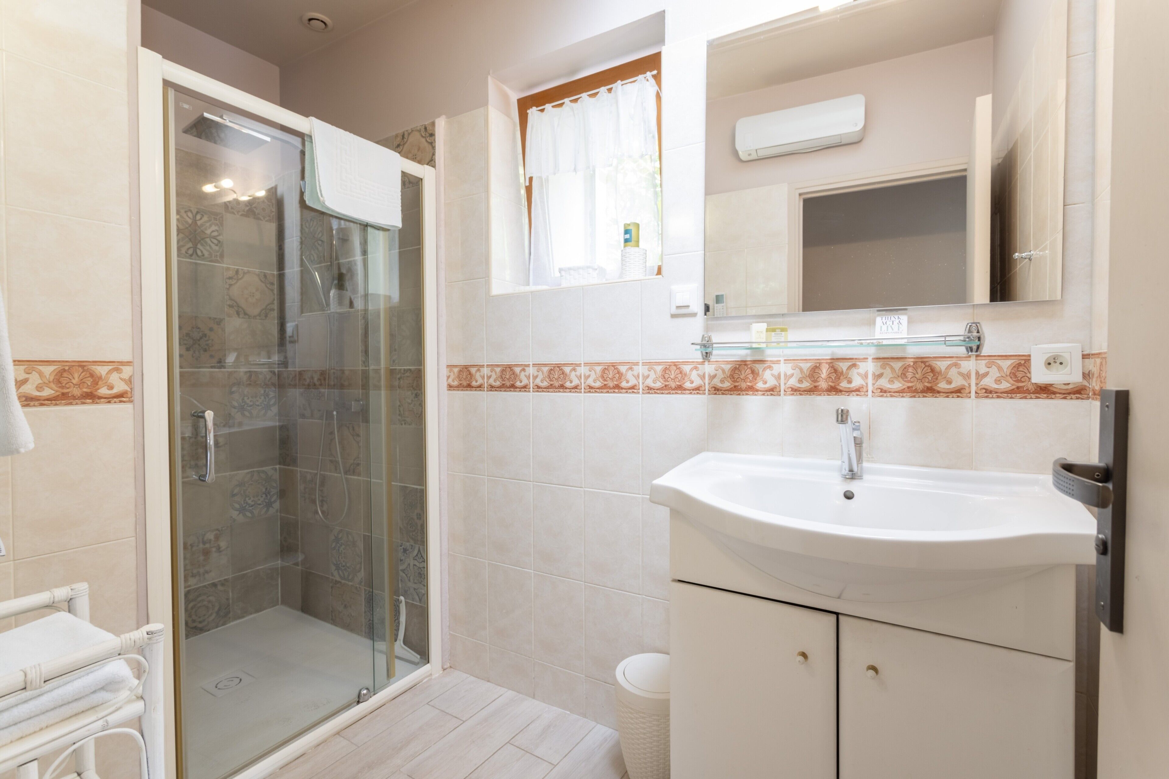 A bathroom with a glass-enclosed shower, a sink, a mirror, and a window with a curtain.