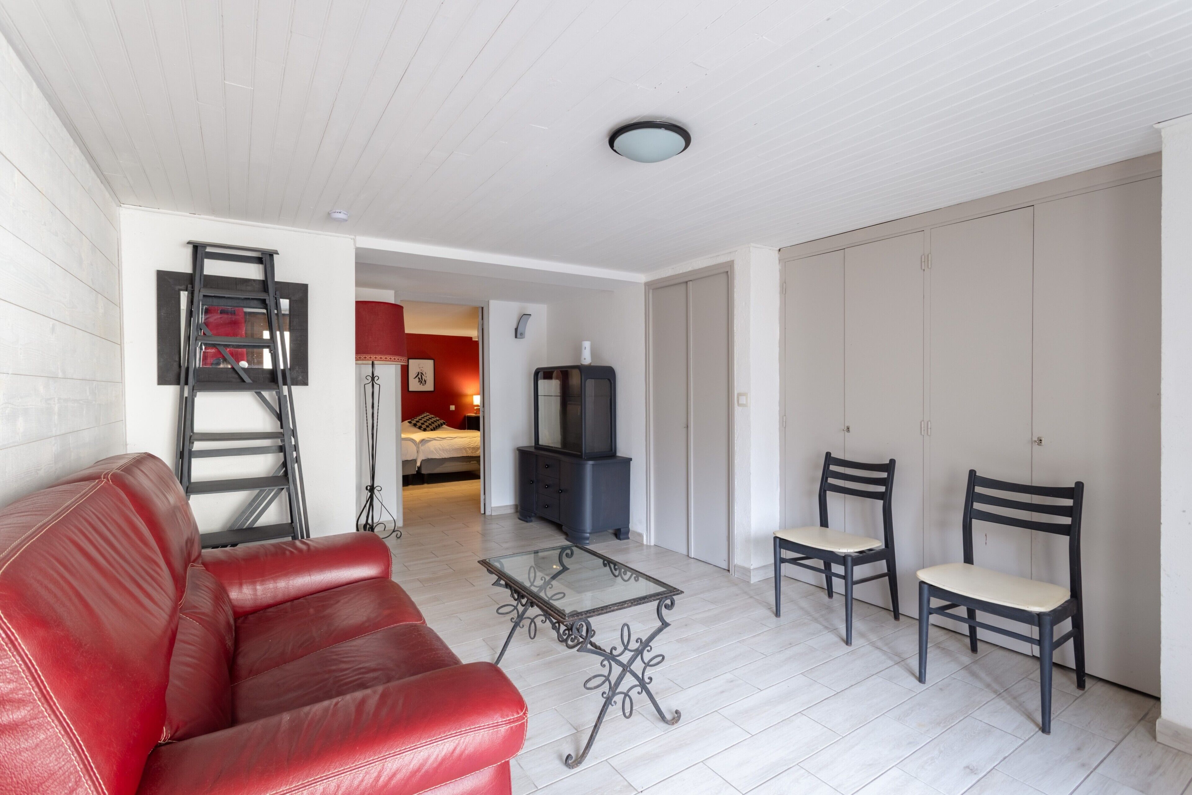 A living room with a red leather sofa, a glass coffee table, two dark wooden chairs, a television, and a wardrobe.