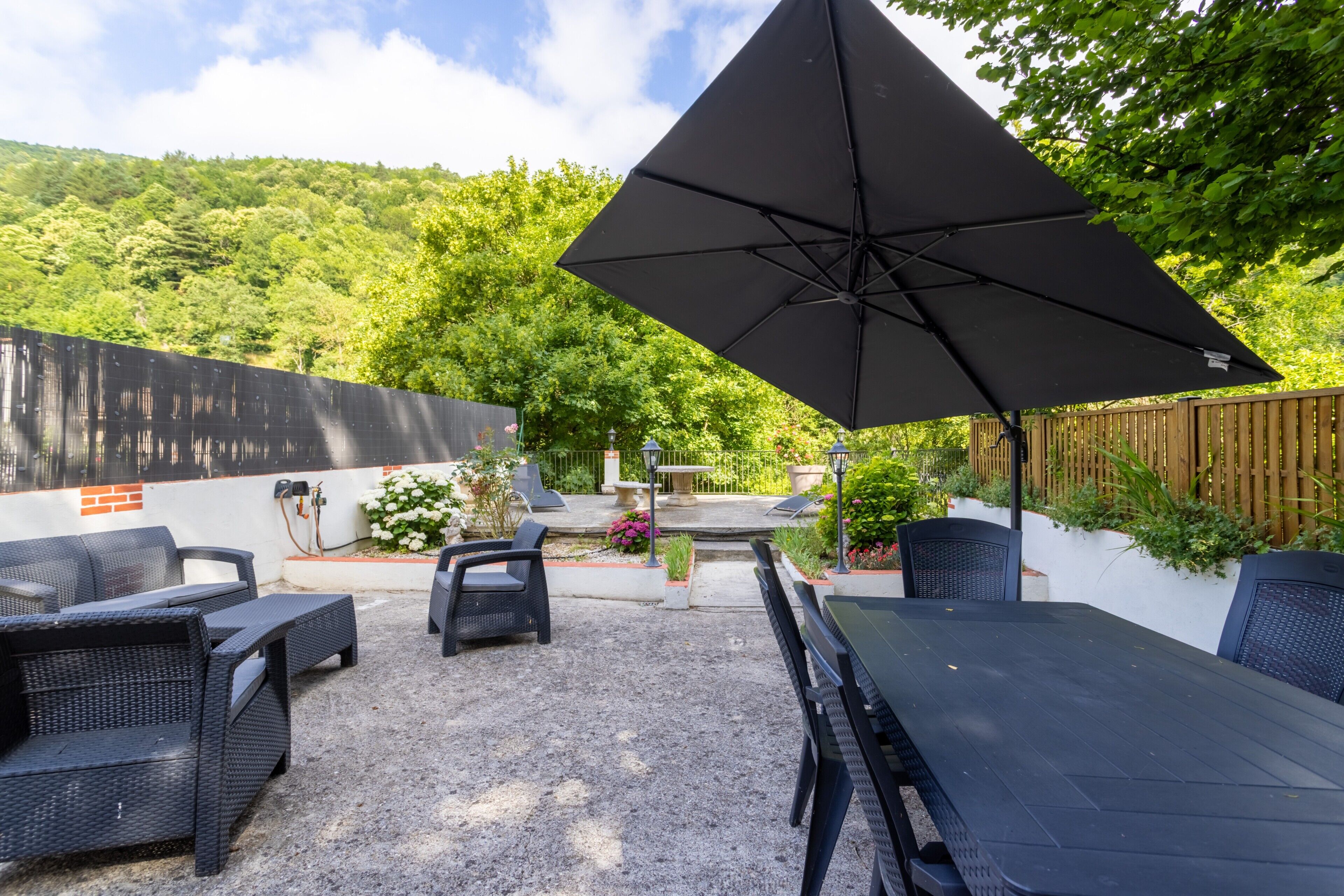 Eine Terrasse im Freien mit Tisch und Stühlen, einem großen Sonnenschirm und Blick auf einen Garten und Hügel.