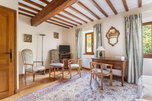 A room with wooden beams, a desk, a chair, a television, and a window with curtains.