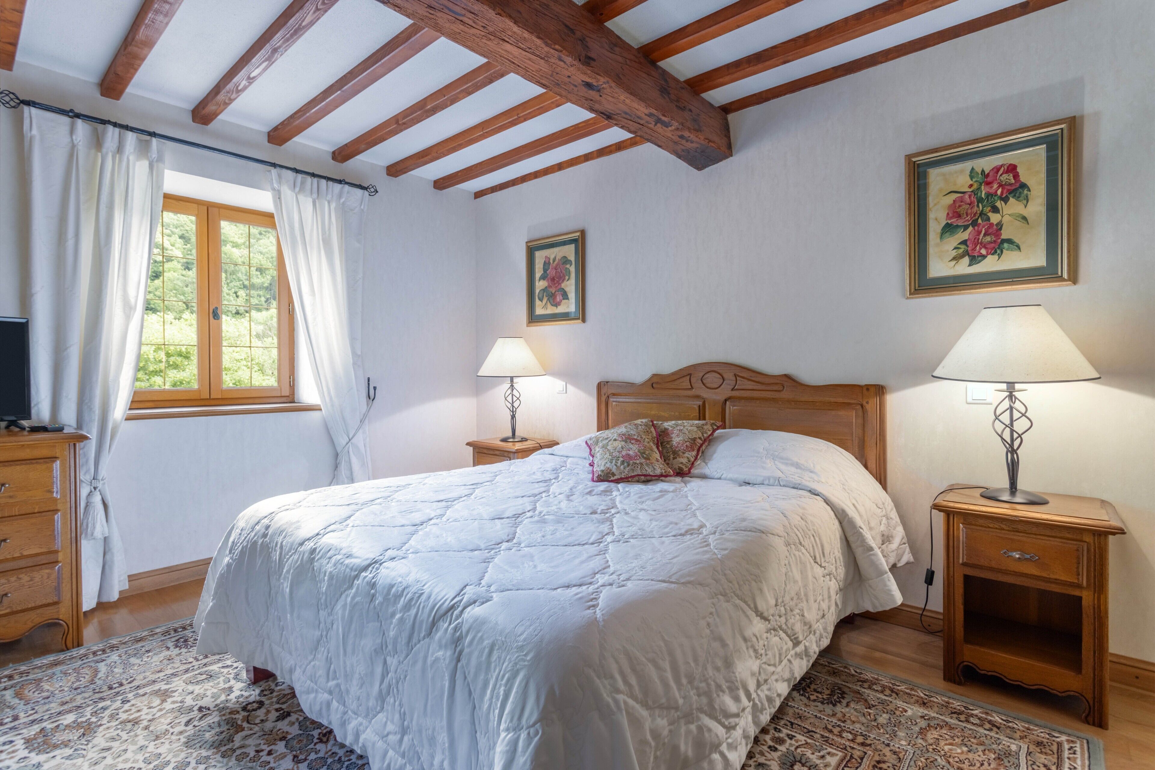 Une chambre avec un plafond en bois, un lit recouvert d’une couette blanche, deux lampes de chevet, une table de chevet, un téléviseur et une fenêtre avec des rideaux.