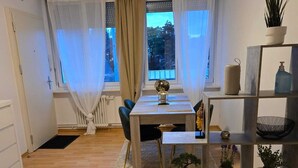 A modern living room with a wooden floor, a dining table with chairs, a sofa, a bookshelf, and a window with curtains.