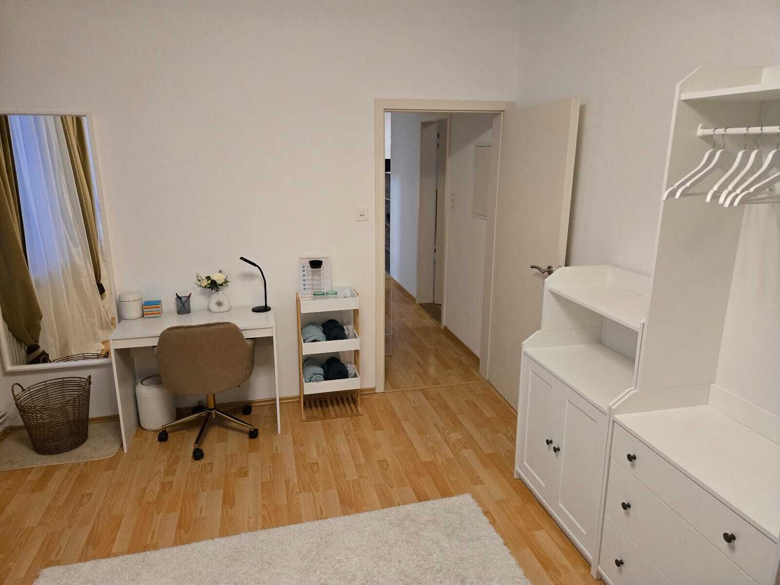 A room with a desk, chair, wardrobe, and a small shelf with items.