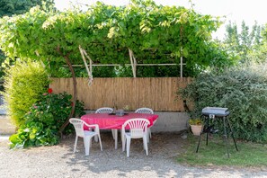 Ein überdachter Essbereich im Freien mit Tisch und Stühlen, einem Grill und viel Grün im Hintergrund.