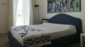 A bedroom with a bed, a window with curtains, a painting on the wall, and a floor lamp. - ‘Vista Castello - Otranto’ Apartment with Sea View, Wi-Fi, and Air Conditioning (Otranto)