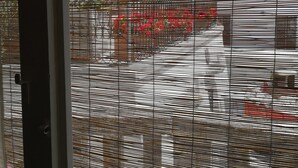 A window with horizontal blinds, a view of a building with a red fence, and a tree. - ‘Vista Castello - Otranto’ Apartment with Sea View, Wi-Fi, and Air Conditioning (Otranto)