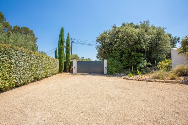 A gravel driveway leading to a gated entrance with a hedge on one side and trees on the other.