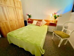 A room with a bed, a chair, a wooden wardrobe, and a nightstand with a lamp and flowers.