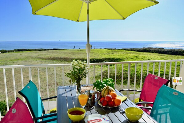 Una mesa con un paraguas amarillo, una jarra, un bol, un plato con fruta y un florero con flores en un balcón con vistas al mar.