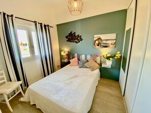 Un dormitorio con cama, una silla, una mesita de noche, una lámpara de pared y una ventana con cortinas.