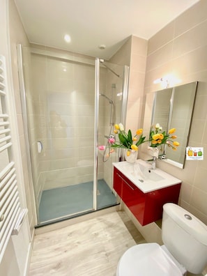 Un baño moderno con cabina de ducha de vidrio, inodoro blanco, tocador rojo y espejo con un florero.
