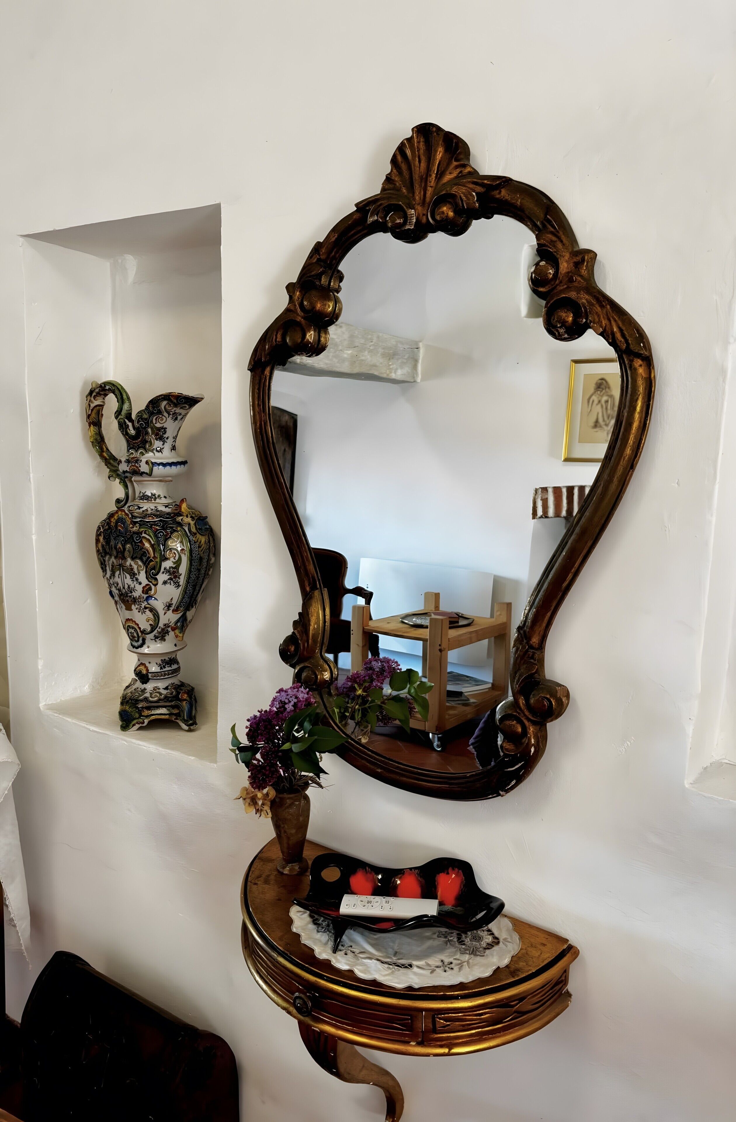 Un miroir mural doté d’un cadre orné, un vase décoratif et une petite table surmontée d’un chemin de table en dentelle.