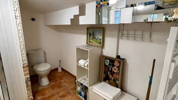 A bathroom with a toilet, shelves with toiletries, a painting, and a mop.