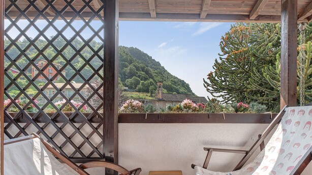 A covered patio with two wooden lounge chairs, a small wooden table, and a view of a mountainous landscape through a lattice screen.