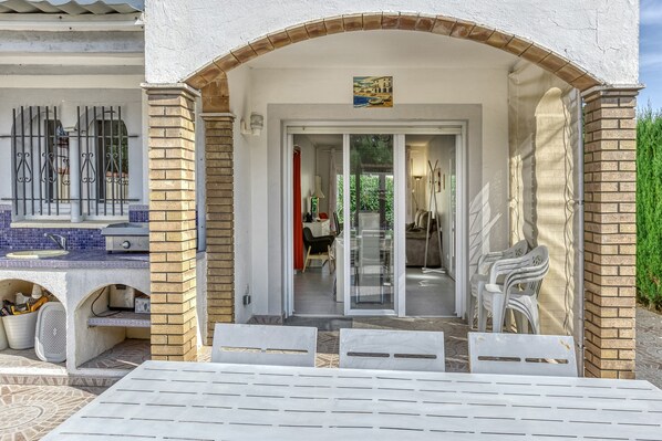 An outdoor patio area with a table and chairs, a kitchen area with a sink and a grill, and a sliding glass door leading to an interior space with a chair and a table.