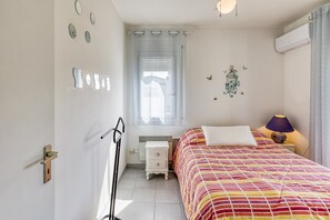 A bedroom with a bed, a nightstand, a radiator, a window with curtains, wall decorations, and an air conditioning unit.
