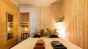 A wooden-paneled room with a bed, towels, a mirror, and a small table with a lamp.