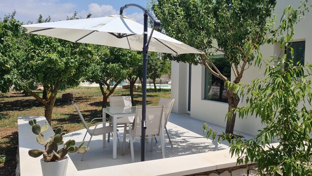A patio with a table and chairs under a large umbrella, surrounded by trees and a white building. - Apartment in semi-detached villa nestled in the Ragusa countryside (Ragusa)