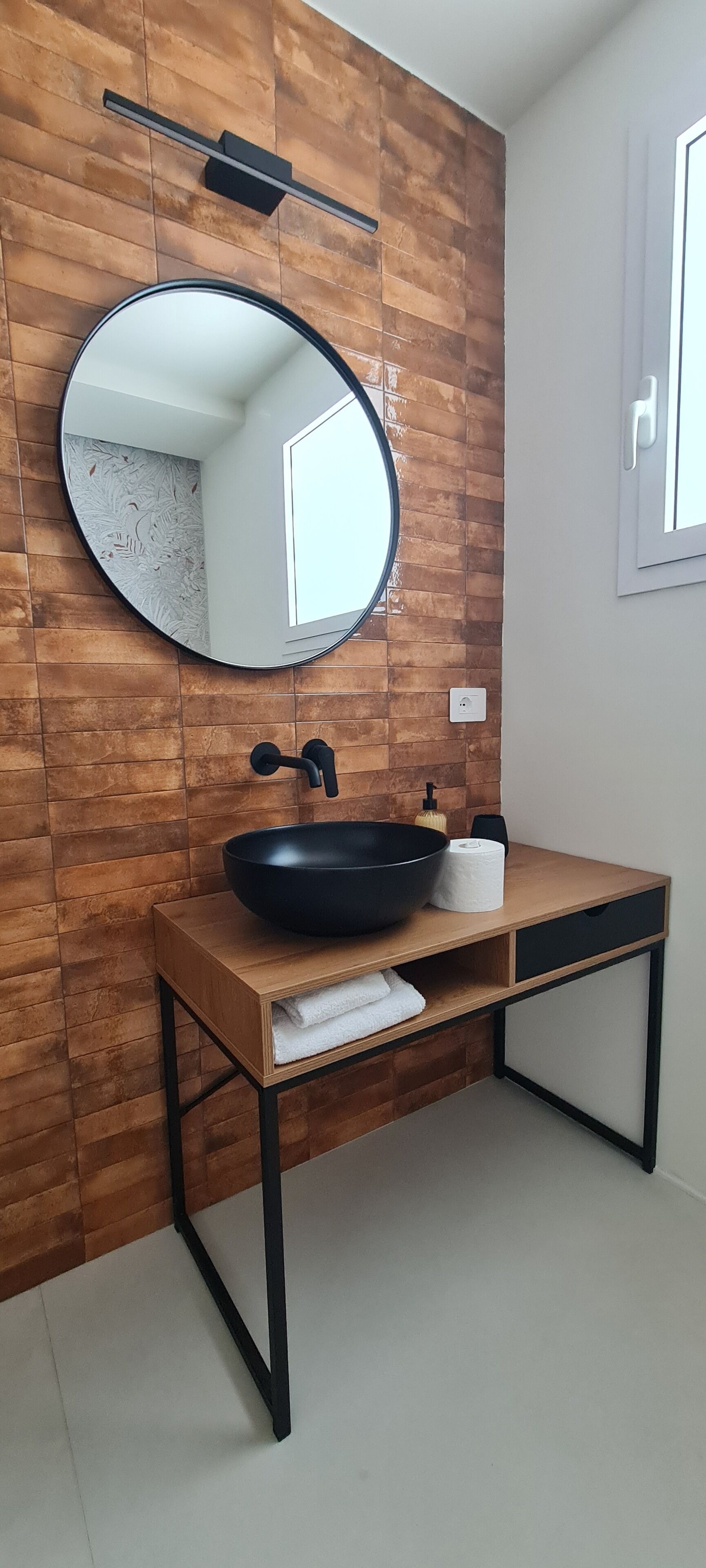 Baño con pared de madera, espejo redondo, lavamanos negro, toalla y dispensador de papel higiénico.