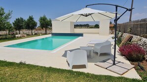 A swimming pool with a sun umbrella, white lounge chairs, and a stone wall. - Apartment in semi-detached villa nestled in the Ragusa countryside (Ragusa)